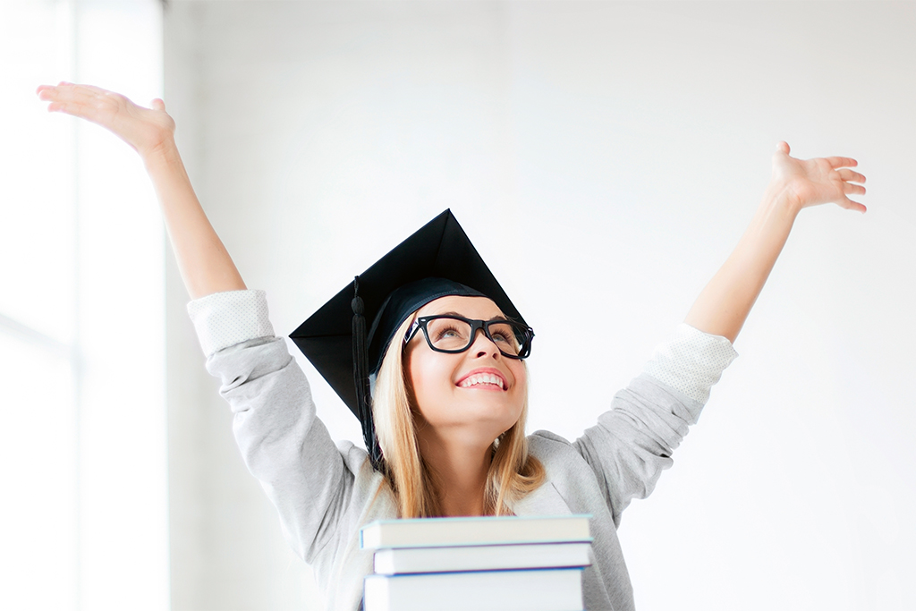 uśmiechnięta studentka w birecie z podniesionymi w górę rękami, na pierwszym planie książki ułożone w stos