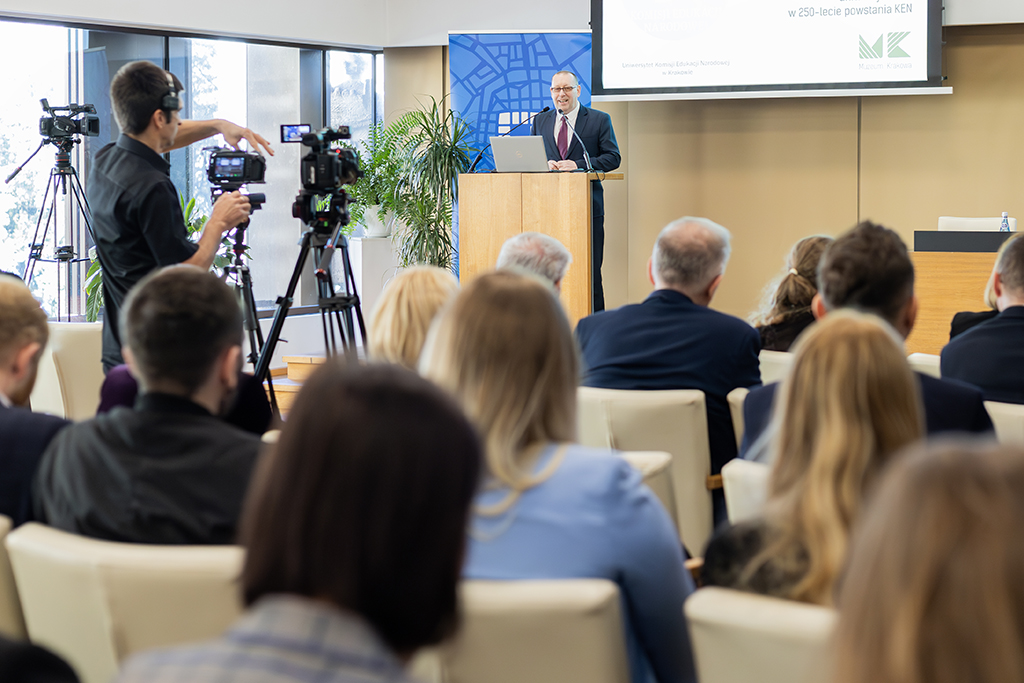 Rektor Uniwersytetu Komisji Edukacji Narodowej w Krakowie prof. dr hab. Piotr Borek przy mównicy, na pierwszym planie uczestnicy spotkania, kamerzysta i 3 kamery